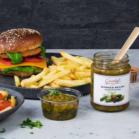 Ein Glas Gurkenrelish von Grashoff neben einem Teller mit einem Burger und Pommes Frites