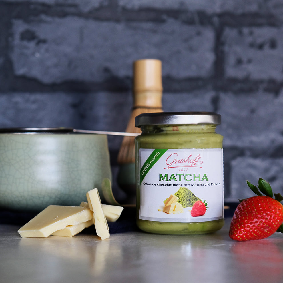 Ein Glas weiße Schokoladencreme mit Matcha und Erdbeeren von Grashoff. Daneben Bruchstücke von weißer Schokolade und eine Erdbeere. Im Hintergrund ein Matcha-Set.