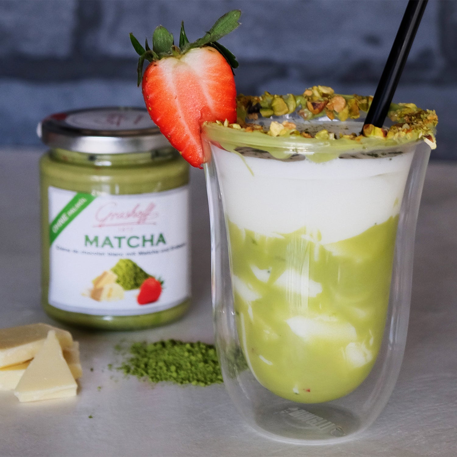 Matcha Eisschokolade im Glas, dekoriert mit einer Erdbeere. Im Hintergrund weiße Schokoladencreme mit Matcha und Erdbeeren von Grashoff.