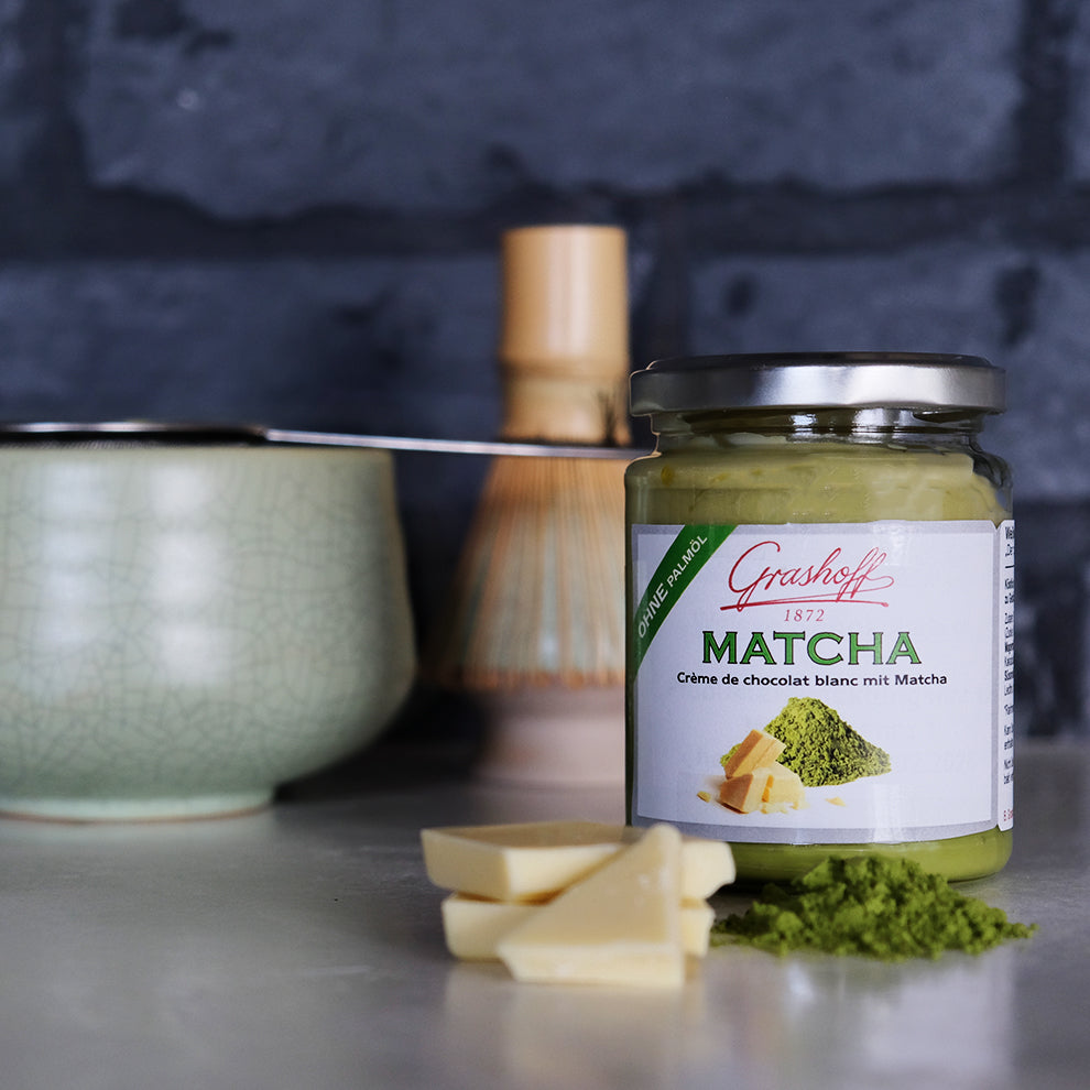 Ein Glas weiße Schokoladencreme mit Matcha von Grashoff. Daneben Bruchstücke von weißer Schokolade und Matcha Pulver Im Hintergrund ein Matcha-Set.